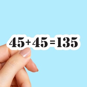 May include: A white sticker with the equation "45 + 45 = 135" in black stencil font. The sticker is held against a light blue background. The numbers and symbols are bold and clear.