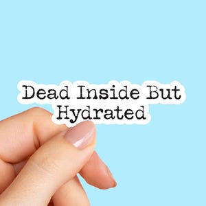 May include: A white sticker with the text "Dead Inside But Hydrated" in a typewriter font. The sticker is held by a hand with a light blue background.