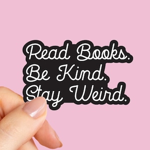 May include: A black sticker with white cursive text that reads "Read Books. Be Kind. Stay Weird." The sticker is held by a hand against a pink background. The sticker has a white border.