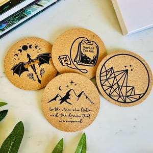 May include: Four round cork coasters with black illustrations. One features a dragon, another a tea bag with "Suriel Tea Co.", a third has mountains and text, and the last has a geometric design. The coasters are on a white surface.