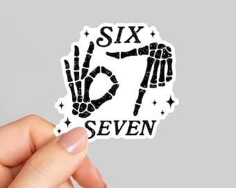 May include: A white sticker with black text and illustrations. The text reads "SIX" above a hand making an "okay" gesture and a hand pointing down, with the text "SEVEN" below. The illustrations are of skeleton hands.