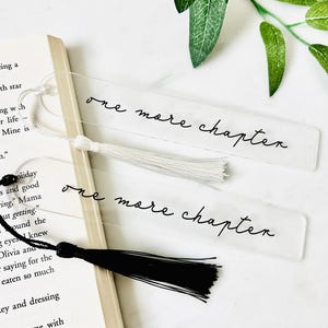 May include: Two clear acrylic bookmarks with the phrase "one more chapter" in black script. One bookmark has a white tassel, the other a black tassel. A book is partially visible in the background.
