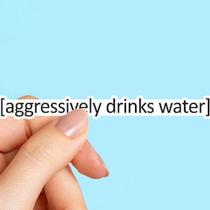 May include: A white sticker with the text "[aggressively drinks water]" is held by a hand with painted nails against a light blue background. The sticker has a black border and the text is in a sans-serif font.