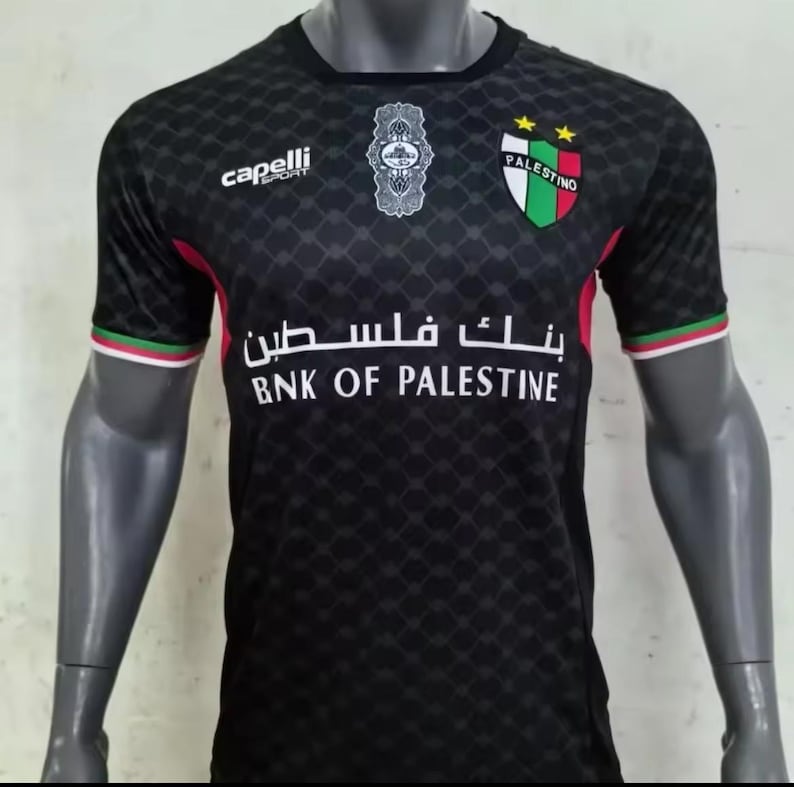May include: Black soccer jersey with a white and green logo that says "Palestino" and a white sponsor logo that says "BNK of Palestine". The jersey has a black and white geometric pattern.