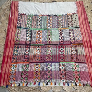 May include: A colorful textile with a white top and a patchwork design. The patchwork features geometric patterns in various colors, including red, green, orange, and purple. The edges are trimmed with red stripes and tassels.