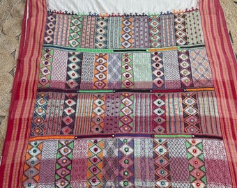May include: A colorful textile with a white top and a patchwork design. The patchwork features geometric patterns in various colors, including red, green, orange, and purple. The edges are trimmed with red stripes and tassels.