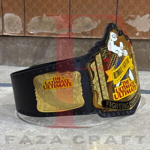 May include: A black and gold championship belt with the text "THE ULTIMATE ULTIMATE FIGHTING CHAMPIONSHIP" on the front. The belt has a gold buckle and a black leather strap. The belt is designed for a fighting competition.