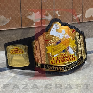 May include: A black and gold Ultimate Fighting Championship belt. The belt features a large gold center plate with the words "Ultimate Fighting Championship" and a fighter graphic. The belt also has a smaller gold plate with the words "The Ultimate Ultimate".