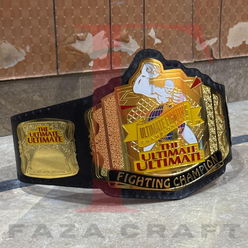 May include: A black and gold Ultimate Fighting Championship belt. The belt features a large gold center plate with the words "Ultimate Fighting Championship" and a fighter graphic. The belt also has a smaller gold plate with the words "The Ultimate Ultimate".
