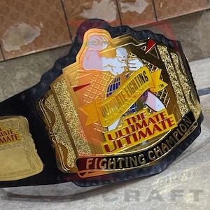 May include: A gold and black Ultimate Fighting Championship belt. The belt features a gold center plate with the words "Ultimate Fighting Championship" and "The Ultimate Fighter Champion". The belt is black with gold accents.