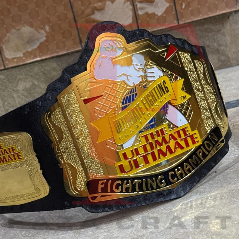 May include: A gold and black Ultimate Fighting Championship belt. The belt features a gold center plate with the words "Ultimate Fighting Championship" and "The Ultimate Fighter Champion". The belt is black with gold accents.
