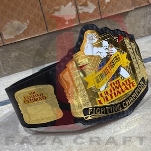 May include: A championship belt with a black leather strap and gold-colored metal plates. The plates feature the text "ULTIMATE FIGHTING CHAMPIONSHIP" and an illustration of two fighters. The belt is a trophy for a fighting competition.