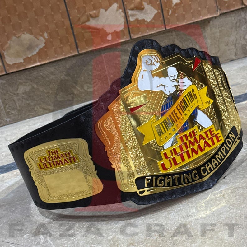 May include: A championship belt with a black leather strap and gold-colored metal plates. The plates feature the text "ULTIMATE FIGHTING CHAMPIONSHIP" and an illustration of two fighters. The belt is a trophy for a fighting competition.
