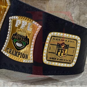 May include: A black leather championship belt with two gold plates. The plates are adorned with rhinestones and the words "FFL Fantasy Football Champion". One plate features a football graphic.