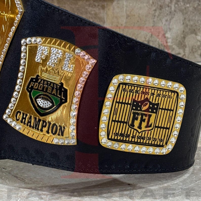 May include: A black leather championship belt with two gold plates. The plates are adorned with rhinestones and the words "FFL Fantasy Football Champion". One plate features a football graphic.