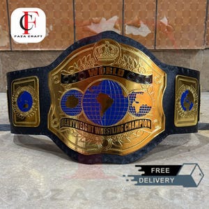 May include: A gold and black heavyweight wrestling championship belt. The belt features a large central plate with the words "WORLD HEAVYWEIGHT WRESTLING CHAMPION" and three globes. The belt has a black leather strap and gold accents. The logo "FAZA CRAFT" is in the top left corner.