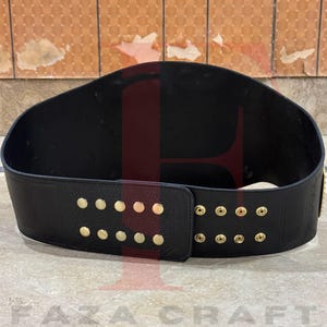 May include: Black leather championship belt with gold studs and accents. The belt has a wide, curved design and a buckle closure. The back of the belt is black. Likely for a wrestling or sports champion.