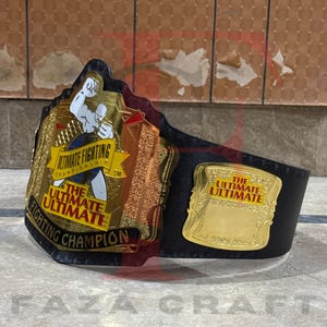 May include: A championship belt with gold and black accents. The belt features the text "ULTIMATE FIGHTING CHAMPIONSHIP" and "THE ULTIMATE ULTIMATE FIGHTING CHAMPION". The belt has a black leather strap.