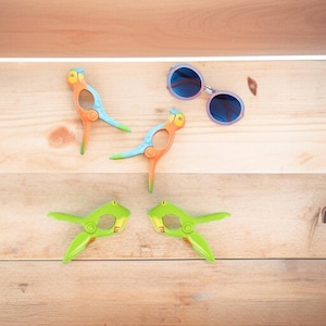 May include: Four colorful plastic clips in the shape of parrots and frogs. The parrot clips are orange, blue, and green. The frog clips are green. The clips are on a wooden surface.