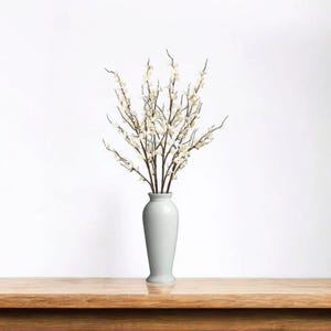 May include: A white ceramic vase holding a bundle of artificial flowering branches. The branches have dark brown stems and small white blossoms. The vase is a light gray color and sits on a wooden surface.