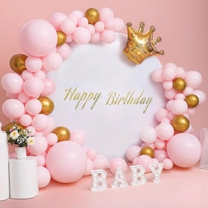 May include: Pink and gold balloon arch with a white round backdrop. The backdrop has the words "Happy Birthday" written on it. There are white letter lights spelling out "BABY" in front of the arch.