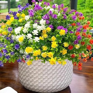 May include: A vibrant arrangement of artificial flowers in a white, textured pot. The flowers are a mix of yellow, purple, white, and orange, with green foliage. The pot has a geometric pattern, creating a modern look. Ideal for home decor.