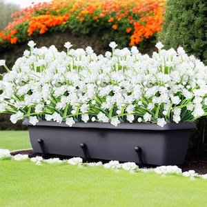 May include: A rectangular, dark gray planter box filled with white flowers and green stems. The planter has three curved hooks on the front. The background features green grass, a hedge, and orange flowers.