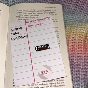 Könnte beinhalten: Ein rosa-weißes "Bookish Enamel Pin" Lesezeichen mit dem Text "Roon Designs" und einer "Constant Reader" Nadel. Das Lesezeichen befindet sich auf einem Buch mit Text auf den Seiten. Der Hintergrund ist ein mehrfarbiger Strickstoff.