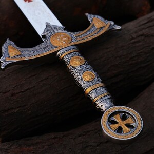 May include: An ornate sword with a silver blade and intricate gold and silver detailing. The hilt features a cross design and decorative patterns. The sword's handle is wrapped in a detailed design. The blade has a red cross.