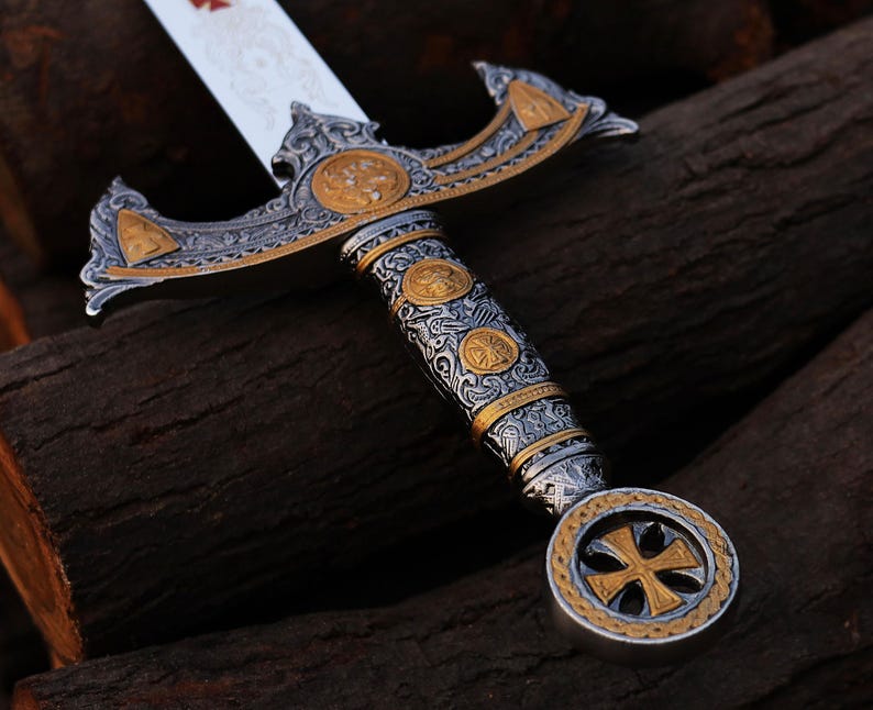 May include: An ornate sword with a silver blade and intricate gold and silver detailing. The hilt features a cross design and decorative patterns. The sword's handle is wrapped in a detailed design. The blade has a red cross.