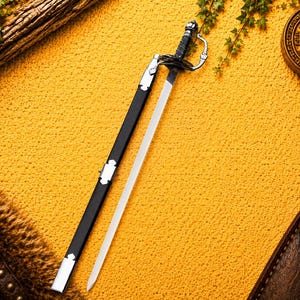 Peut inclure: Une épée décorative avec une lame argentée et une poignée et un fourreau noirs. L'épée est exposée sur une surface jaune texturée, avec une bûche de bois et du feuillage vert en arrière-plan. Un étui en cuir marron est visible.