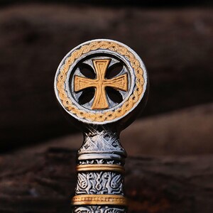 May include: Close-up of a silver and gold-toned cross pendant. The cross is centered within a circular frame with a gold braided border. The pendant has intricate detailing and a textured finish.