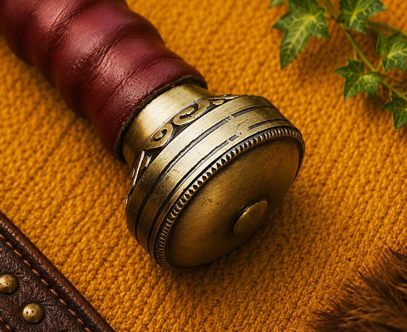 May include: A sword pommel crafted from antique brass featuring intricate engraved scrollwork and a beaded edge detail. The pommel is attached to a handle wrapped in deep burgundy leather, showcasing a metallic finish and textured design.