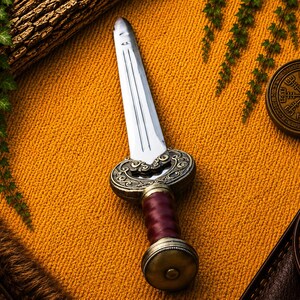 May include: A double-edged dagger with a polished steel blade, a dark red leather-wrapped grip, and an ornate bronze-toned metal crossguard and pommel. The crossguard features intricate scrollwork, and the pommel is a rounded, textured disc.