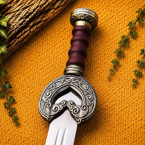 May include: A sword with a polished steel blade and a dark purple, ribbed leather grip. The hilt features an ornate, antiqued brass crossguard with intricate swirling patterns and a matching brass pommel at the top of the handle.