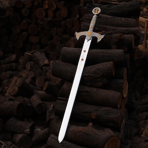 May include: A decorative sword with a silver blade and ornate gold and silver hilt. The hilt features a cross design. The sword is leaning against a stack of dark brown logs.