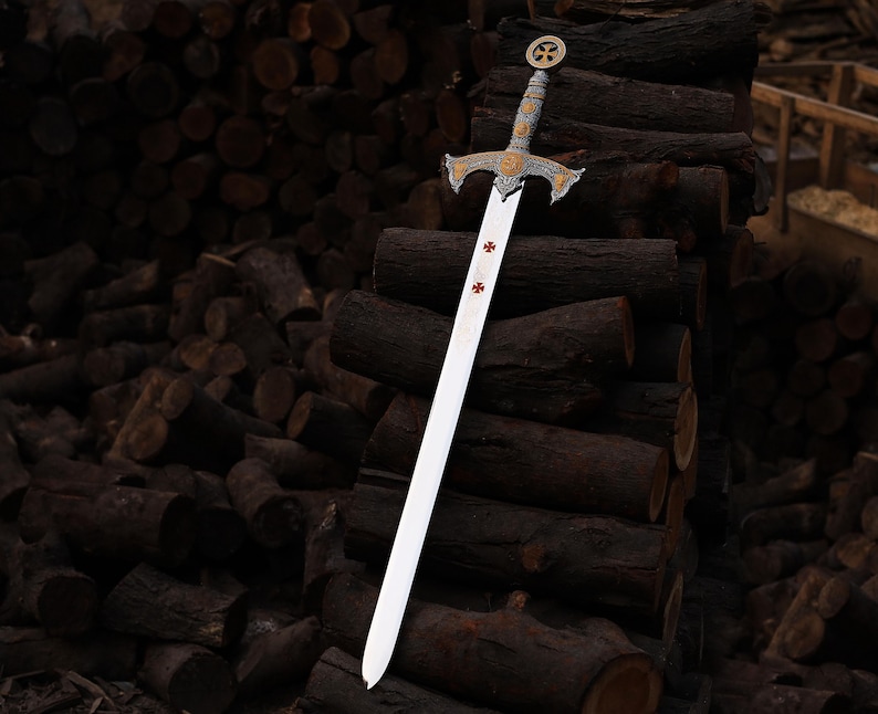 May include: A decorative sword with a silver blade and ornate gold and silver hilt. The hilt features a cross design. The sword is leaning against a stack of dark brown logs.