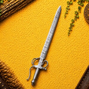May include: A silver-toned metal letter opener featuring a long, textured blade with a wavy pattern and a detailed hilt with curved guards and a rounded pommel. The object is positioned diagonally against a bright yellow textured background.