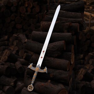 May include: A decorative sword with a silver blade and ornate gold and silver hilt. The blade features red cross symbols. The sword is displayed against a backdrop of stacked logs, showcasing its historical design.