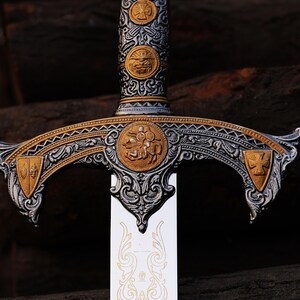 May include: Close-up of an ornate sword with a silver blade and a detailed hilt. The hilt features gold and silver accents, including circular emblems and decorative patterns. The sword's design suggests a historical or fantasy theme.