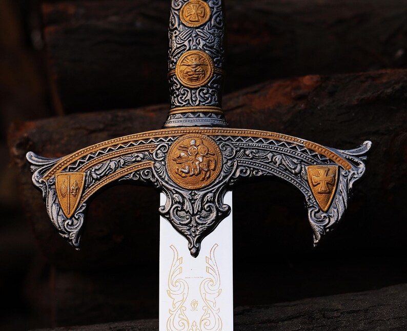 May include: Close-up of an ornate sword with a silver blade and a detailed hilt. The hilt features gold and silver accents, including circular emblems and decorative patterns. The sword's design suggests a historical or fantasy theme.