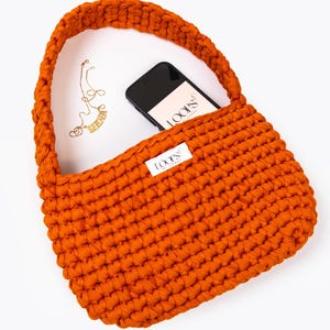 Könnte beinhalten: Eine leuchtend orangefarbene gehäkelte Handtasche mit einem dicken Riemen. Die Tasche hat ein kleines weißes Etikett mit dem Text "LOOPS BY NATASHA" darauf. Eine goldene Kettenkette und ein schwarzes Smartphone befinden sich in der Tasche.