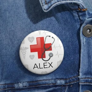 Puede incluir: Un botón blanco con una cruz roja y un diseño de estetoscopio. El botón también presenta el nombre "ALEX" en texto negro, junto con iconos de temática médica. El botón está sobre un fondo de mezclilla azul.