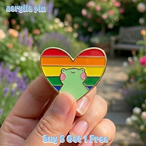 Puede incluir: Pin de acrílico en forma de corazón con diseño de arcoíris e ilustración de rana verde. El pin tiene un borde dorado y el texto "acrylic pin" en la parte superior. El texto "Buy 5 Get 1 Free" está en la parte inferior.