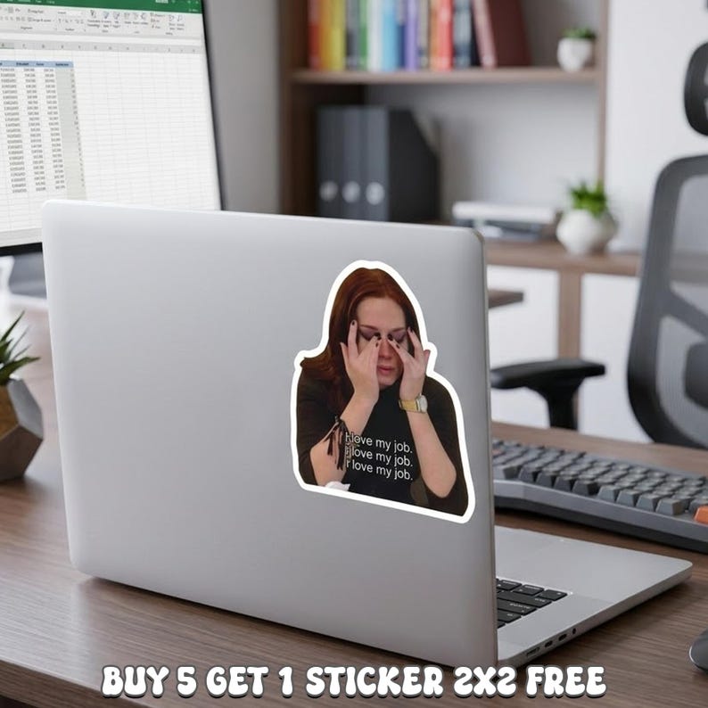 May include: A white sticker on a laptop featuring a woman with red hair, covering her eyes. The sticker has the text "love my job" repeated three times. The laptop is on a wooden desk in an office setting.