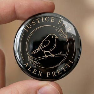 Puede incluir: Un botón negro con texto dorado y una ilustración de un pájaro. El texto dice "JUSTICE FOR ALEX PRETTI" en el borde, con un pájaro en una rama en el centro. El botón está sostenido entre dos dedos.