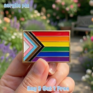 May include: A rectangular acrylic pin with a gold border, featuring the Progress Pride flag design. The flag includes the traditional rainbow stripes, plus the colors of the transgender flag, and black and brown stripes. The text "acrylic pin" is at the top.