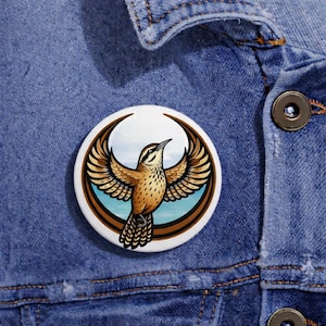May include: A round button pin featuring a detailed illustration of a bird with outstretched wings. The bird is brown and tan, set against a backdrop of blue sky and water, framed by a brown border. The pin is set against a denim jacket.