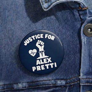 May include: A navy blue button with white text and graphics. The text reads "JUSTICE FOR ALEX PRETTI" with a raised fist, a heartbeat, and a medical symbol. The button is pinned to a denim jacket.