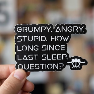 May include: A black sticker with white text that reads "GRUMPY. ANGRY. STUPID. HOW LONG SINCE LAST SLEEP, QUESTION?" The sticker has a rounded rectangular shape and a small white graphic at the bottom right.